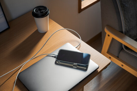Charging smartphone on closed laptop with coffee cup on desk