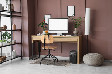 Fototapeta premium Interior of modern office with workplace, shelving unit and floor lamp near brown wall