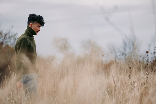 A contemplative young man walks through tall dry grass in muted overcast light, evoking authenticity and truthful emotion with natural posture, simple clothing and quiet presence.