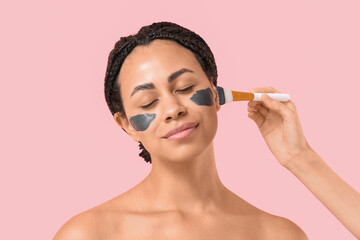 Pretty young African-American woman and hand with brush applying activated charcoal mask on face against pink background