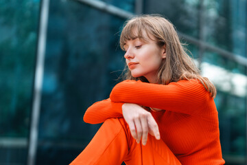 Young woman with long hair wearing orange outfit sitting against glass wall in an urban setting...