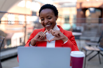 Woman making heart shape with hands while working on laptop at a cafe terrace in the afternoon