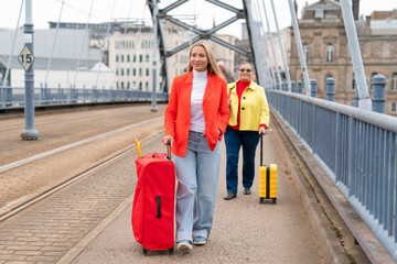 Two women enjoy a spring stroll across a city bridge while pulling their colorful luggage behind...