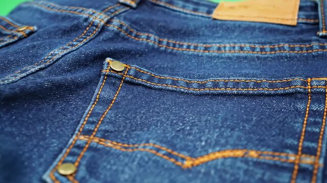 Folded blue jeans with orange stitching and brass button details, set against a green backdrop