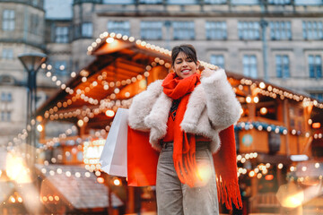 Woman enjoying a festive market in a cozy winter outfit during the holiday season, holding shopping bags and smiling in a decorated square