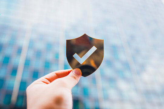 Hand holding shield with check mark icon in front of office building background, representing business security, data protection, trust validation, cybersecurity, and corporate assurance.