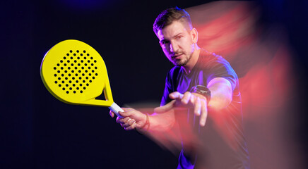 Man playing padel with a yellow racket in a dark setting with colorful lighting effects