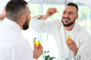 Handsome young man applying vitamin E serum and looking at mirror in bathroom
