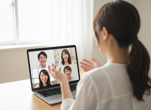 明るい日差しが差し込む部屋で、ノートパソコンの画面に映る4人の日本人男女と笑顔でオンライン会話/コミュニケーションを楽しむ20代の日本人女性