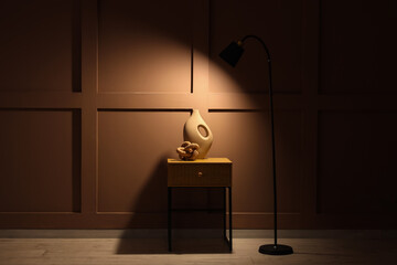 Wooden bedside table with vase and glowing floor lamp near brown wall in room