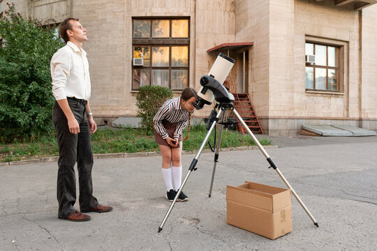A student carefully observes telescope adjustments with her teacher's guidance during an astronomy class outside a building, focusing on precision for sky exploration