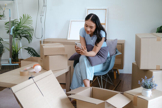 Asian woman using mobile phone among moving boxes for home relocation