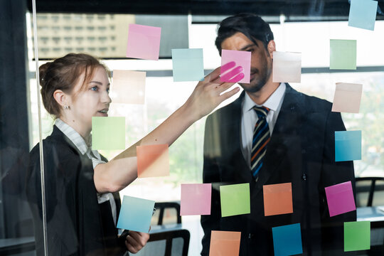 Asian businessman and caucasian businesswoman standing behind glass board discussing colorful sticky notes together during creative brainstorming session in modern office environment - Powered by Adobe