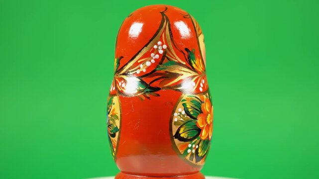 A close-up of a Russian wooden doll, with a red headscarf and floral decor, on green backdrop