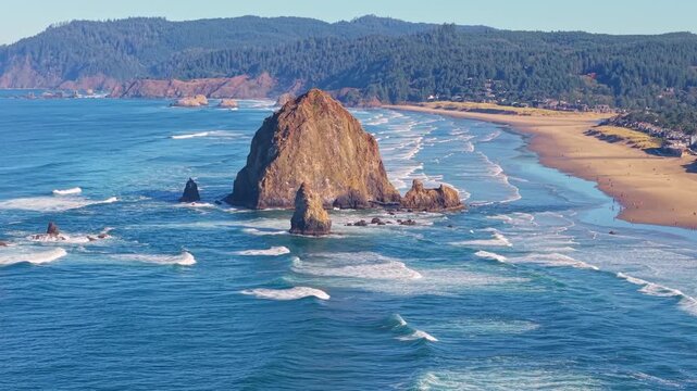 A breathtaking high-angle video capturing the iconic Haystack Rock sea stack, the expansive sandy beach, and the Pacific Ocean coastline under a clear, bright, sunny sky at Cannon Beach, Oregon