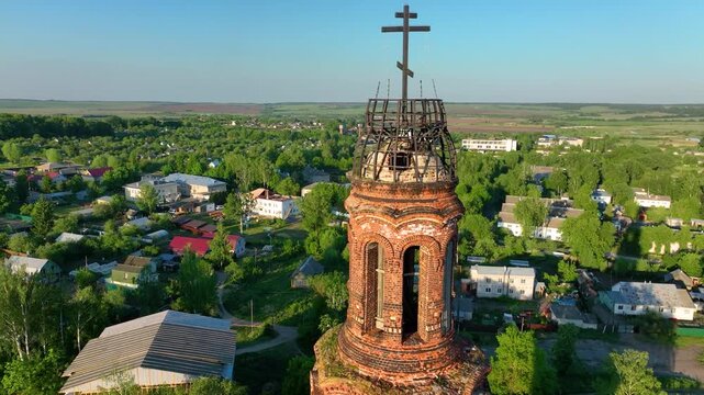 Close up aerial view of the upper section of a historical, weathered red brick bell tower towering over the green rural townscape