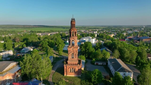 Close up aerial view of the upper section of a historical, weathered red brick bell tower towering over the green rural townscape