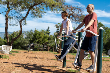 A middle aged Caucasian couple is exercising  using outdoor fitness equipment outdoors at a public park.