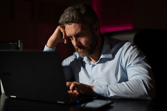 Business man working overtime in a dark office late at night. Overworked employee with laptop in night office. Freelancer concentrating on work. Worker sitting at a desk looking tired and bored.