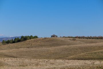 A solitary wooden cabin sits on a gentle hill in a wide open field. A line of pine trees and distant hills frame the scene beneath a bright, calm blue sky.