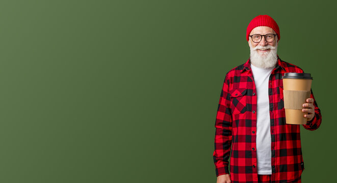 Elderly man dressed as Santa Claus holding takeaway coffee cup in red plaid shirt and hat. Festive holiday portrait for winter celebrations. Christmas and New Year delivery concept with jolly vibe.