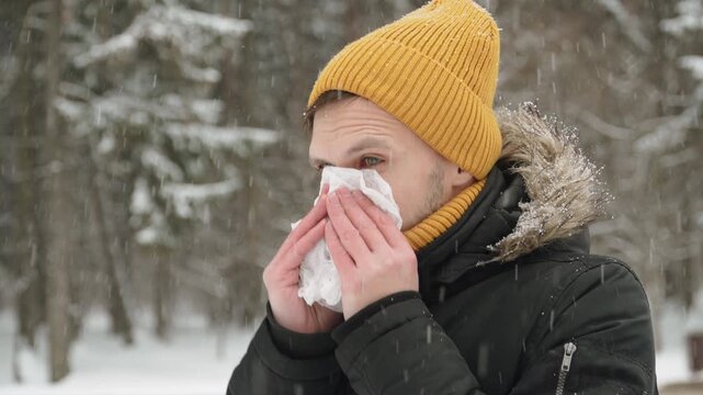 Caucasian man blows his nose during snowfall in city park with trees. Portrait of person suffering from seasonal viral diseases in winter.