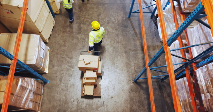 Logistics, man or walking with trolley in warehouse to transport boxes, supply chain or shipping. Above, distribution picker and moving packages for inventory export, storage management and delivery