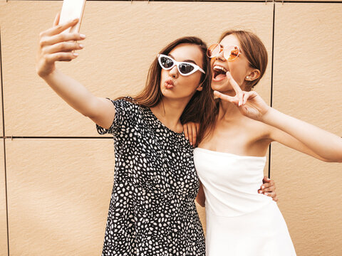 Two young smiling hipster women in summer clothes. female taking selfie self portrait photos on smartphone.Models posing on street background near yellow wall.Female showing positive face emotions