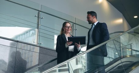 Smiling colleagues discussing financial data on tablet while going down modern escalator in office building - Powered by Adobe