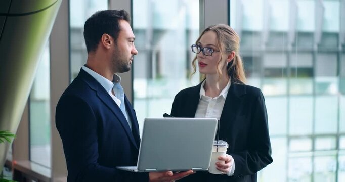 Male and female professionals collaborating over a laptop computer while standing together by large windows in a bright modern corporate setting