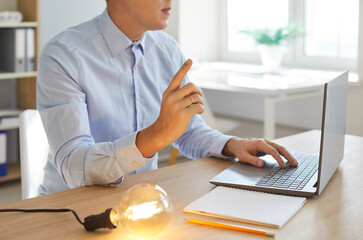 Young intelligent businessman with laptop, paper and glowing light bulb changes marketing plan...