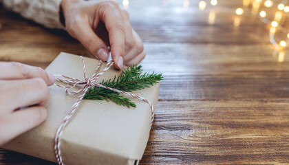 Hands wrapping a festive gift with string and an evergreen branch on a wooden table with blurred lights