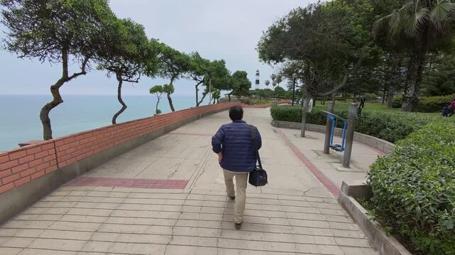 Camera follows TV host Charles Huang walking Malec&oacute;n de Miraflores boardwalk toward La Marina Lighthouse, with Pacific Ocean cliffs on left and bench seats on right, surrounded by trees
