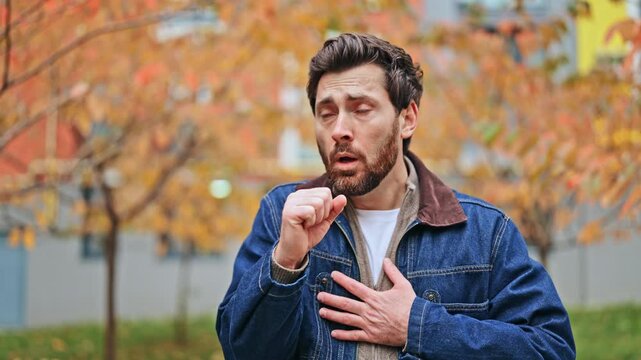Unwell man experiences persistent cough outdoors, displaying significant discomfort and pain. Symptoms suggest seasonal cold, flu, or respiratory illness amidst autumn foliage.