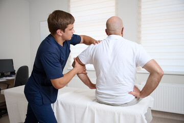 Obraz premium Chiropractor adjusting male patient’s shoulder in clinic. Physiotherapist performing manual therapy to relieve pain, improve mobility, support rehabilitation after injury or musculoskeletal disorder.