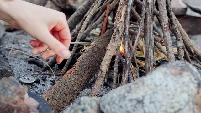 Hand arranging driftwood teepee over embers in a campfire ring