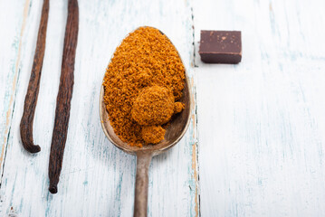 pile of brown sugar on old spoon, vanilla, broken dark chocolate blocks on white wooden table