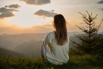 Obraz premium Woman sits peacefully on a grassy hill, gazing at the sunset as golden light bathes the landscape. Rolling mountains and a single evergreen tree create a tranquil atmosphere in the evening.