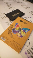 Naklejka premium A 'Happy Color' drawing notebook adorned with a vibrant butterfly illustration rests on a table alongside a 'Harry Potter' branded pouch, hinting at a casual moment of artistic creativity