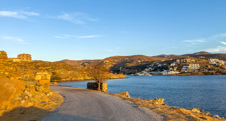 Scenic coastal road at sunset overlooking the calm bay of Vourkari within Kea, Tzia, Greece, featuring golden light upon dry hills, stone walls, and seaside houses under the clear blue sky
