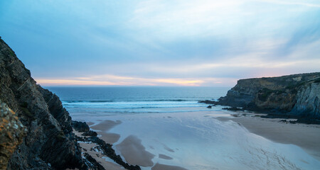 The serene beach at sunset features gentle waves kissing the sandy shore, surrounded by rugged cliffs on Portugal's coastline. The sky's pastel hues add a calming ambiance, perfect for a tranquil.