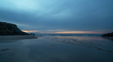 The serene beach at sunset features gentle waves kissing the sandy shore, surrounded by rugged cliffs on Portugal's coastline. The sky's pastel hues add a calming ambiance, perfect for a tranquil.