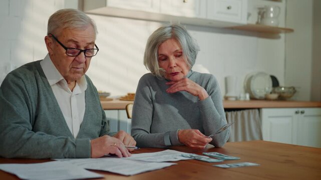 Senior spouses spending weekend at home, counting money and planning expenses. Portrait of elderly man and woman in kitchen home interior, mandatory payments and savings for future, caucasian couple
