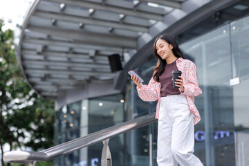 Fototapeta premium Happy young asian woman walking, using smartphone, holding coffee