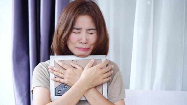 Grieving Asian woman crying while holding a photo of a loved one who passed away, expressing deep sadness, loneliness, and emotional pain. Concept of mourning, loss, heartbreak, and emotional healing