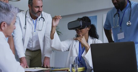 Lab coat doctor wearing VR headset gesturing in hospital conference room, with laptop, stethoscopes