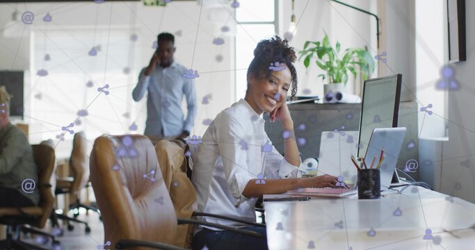 Typing female professional wearing business attire at office desk with laptop, plant, network icons