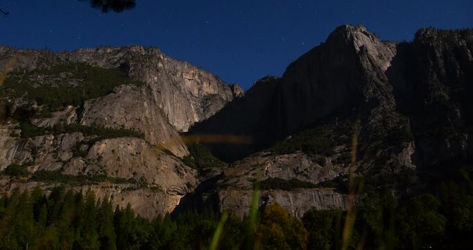 A stunning night timelapse of Yosemite Falls illuminated by moonlight beneath a clear starry sky. Granite cliffs, forested valley, and flowing water create a peaceful natural atmosphere.