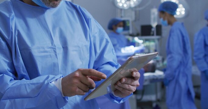Reviewing data on tablet, surgeon in scrubs guiding team in operating room, with monitors and trays - Powered by Adobe