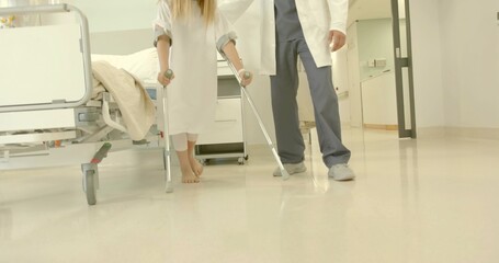 Walking kid wearing hospital gown using forearm crutches along hospital corridor, with doctor help
